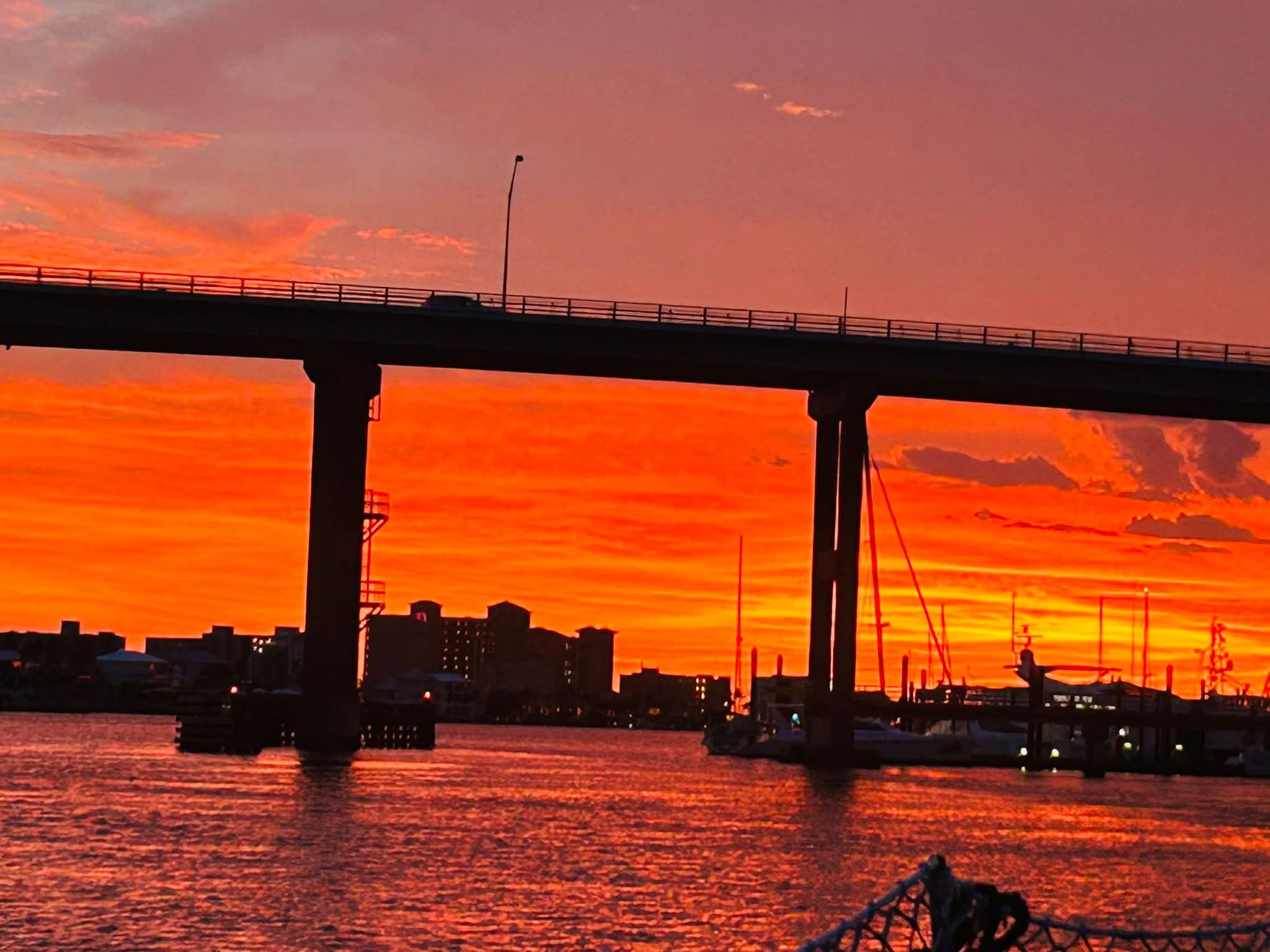 Vibrant orange sunset behind a silhouetted bridge and city skyline over reflecting water.