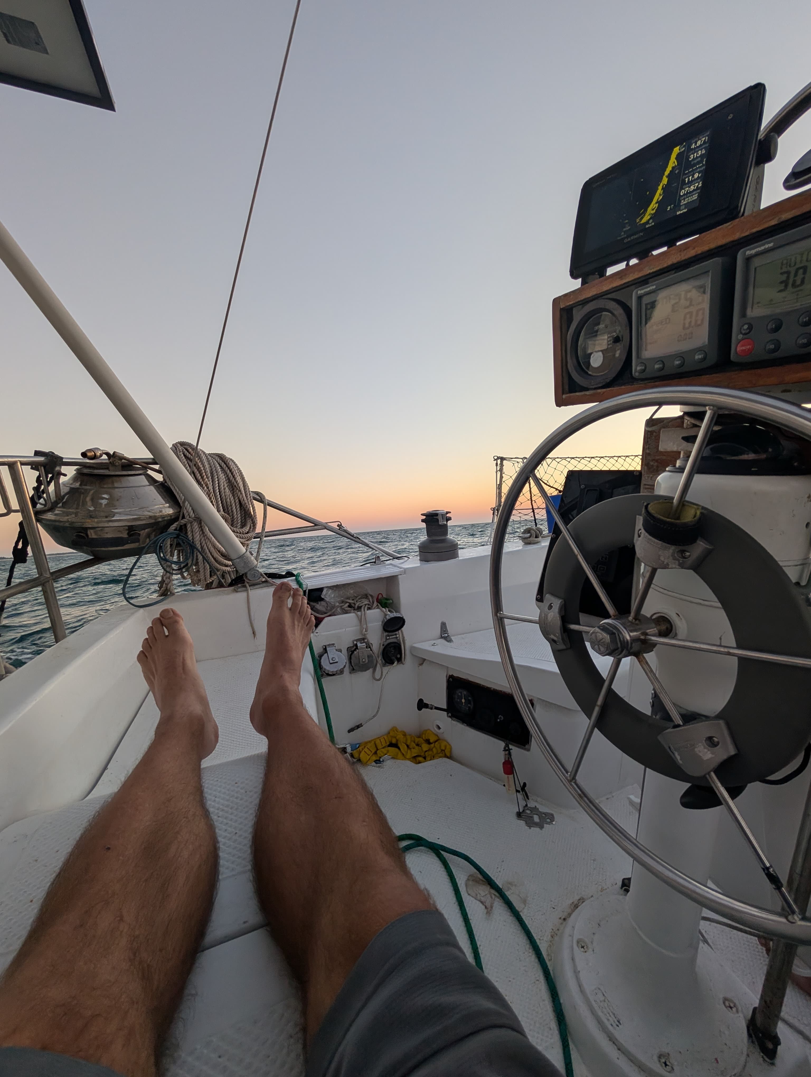 Bare legs on a sailboat deck at sunset, with the helm and ocean horizon.