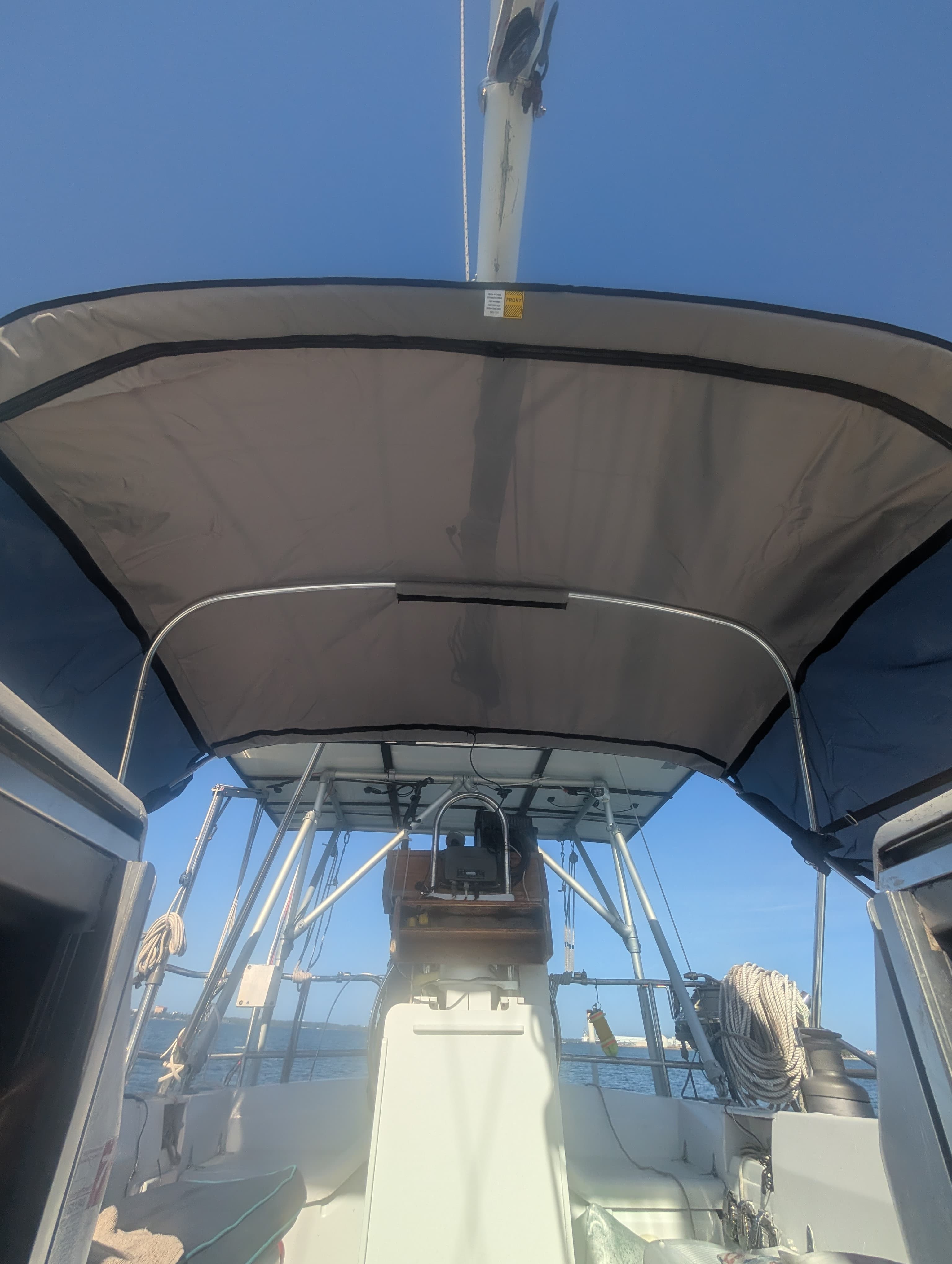 View from a sailboat cockpit under a grey bimini top with steering wheel and rigging.